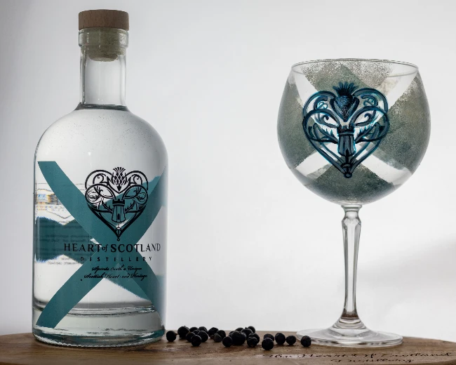 Heart of Scotland Gin bottle with a hand-painted glass on an oak board, surrounded by juniper berries, highlighting premium Scottish craft distillation.
